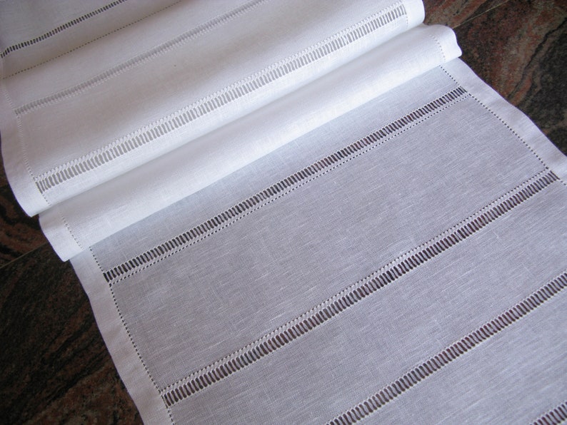 May include: White linen table runner with a delicate stitched design. The runner has a subtle texture and a simple, elegant look.