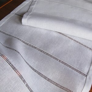 May include: White linen table runner with a delicate embroidered design. The runner has a subtle sheen and a soft texture.