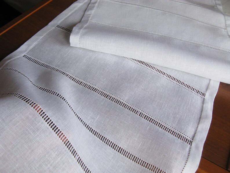 May include: White linen table runner with a delicate embroidered design. The runner has a subtle sheen and a soft texture.
