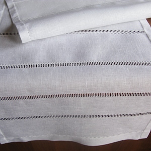 May include: White linen table runner with delicate stitched lines. The runner is folded in half, showcasing the intricate stitching detail.