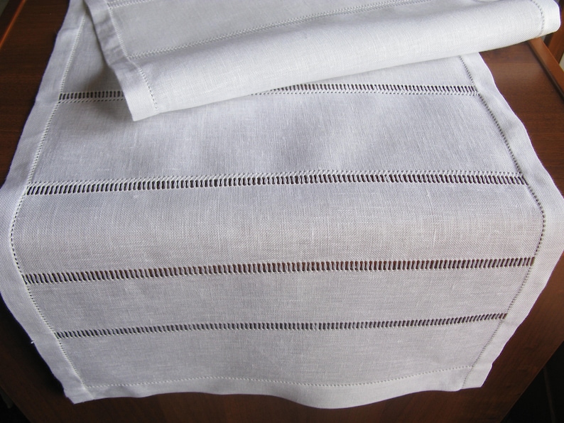 May include: White linen table runner with delicate stitched lines. The runner is folded in half, showcasing the intricate stitching detail.