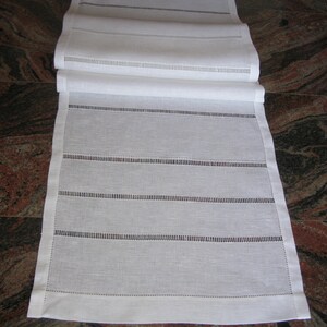 May include: White linen table runner with a delicate embroidered design. The runner features a series of horizontal lines with small, evenly spaced stitches.