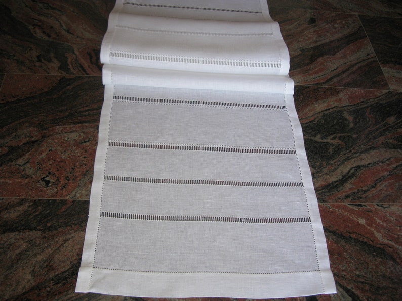 May include: White linen table runner with a delicate embroidered design. The runner features a series of horizontal lines with small, evenly spaced stitches.