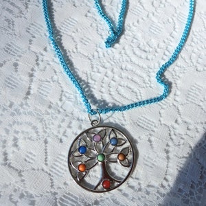 Puede incluir: Un collar con un colgante de plata en forma de árbol de la vida con piedras preciosas de colores. El collar está en una cadena azul claro.