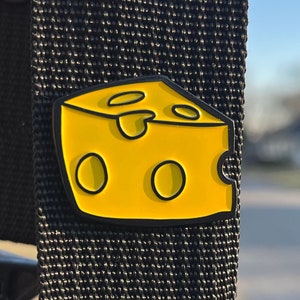 Puede incluir: Un pin de esmalte amarillo con forma de bloque de queso con agujeros. El pin tiene un contorno negro y está sujeto a un fondo de tela negro.