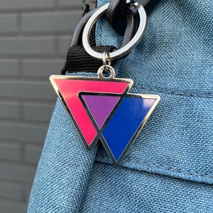 Könnte beinhalten: Ein silberner Schlüsselanhänger mit einem pinkfarbenen, violetten und blauen Dreiecksdesign. Der Schlüsselanhänger ist an einem blauen Rucksack befestigt.