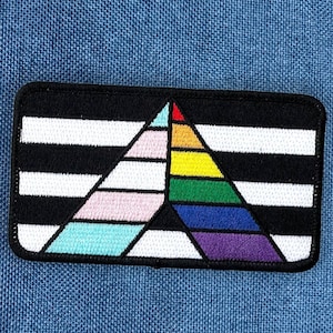 May include: A rectangular patch with a black border and a geometric design. The design features a black and white striped background with a colorful triangle composed of rainbow colors, pink, and turquoise. The patch is on a blue fabric.