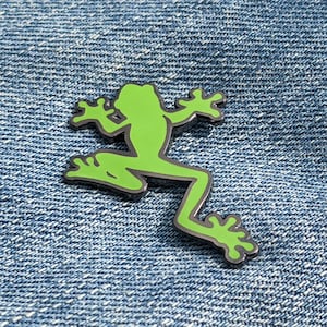 Peut inclure: Épinglette en émail en forme de silhouette de grenouille verte sur un fond en jean bleu.