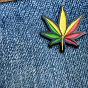 Pode incluir: Um broche de esmalte verde, amarelo e vermelho em forma de folha de marijuana. O broche está sobre um fundo de jeans azul.