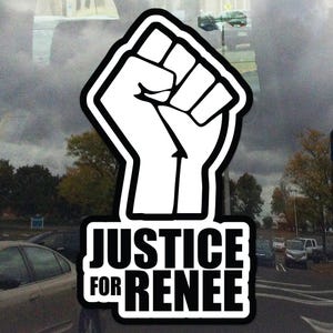 May include: A black and white sticker featuring a raised fist symbol with a thick black outline. The text "JUSTICE FOR RENEE" is printed in bold, black capital letters below the fist. The sticker is placed on a window with a blurred outdoor background.