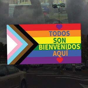 May include: A rectangular sticker with a rainbow flag design and the words "TODOS SON BIENVENIDOS AQUÍ" in green. The flag includes a triangle with the colors of the transgender flag and a brown and black triangle. A purple heart is above and a red heart is below the text.