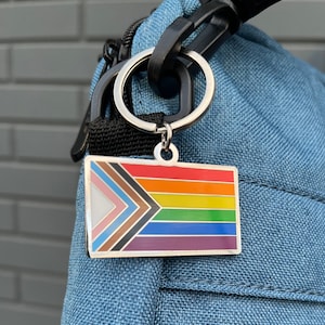 Op de afbeelding: Een zilveren sleutelhanger met een regenboog-pride-vlagcharme. De vlag heeft zes strepen: zwart, bruin, lichtroze, wit, lichtblauw en lichtgeel. De charme is bevestigd aan een zilveren sleutelring.