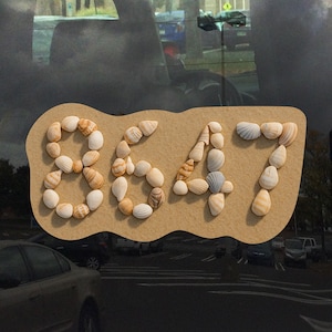 Puede incluir: Un letrero decorativo con el número 8647, hecho con conchas marinas. Los números están dispuestos sobre un fondo beige con bordes ondulados. Las conchas marinas son de varias formas y colores, creando un diseño único con temática de playa.