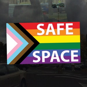 May include: A sticker with a rainbow flag and the words "SAFE SPACE" in white letters. The flag has the colors red, orange, yellow, green, blue, indigo, and violet. The flag also has a black, brown, light blue, and pink triangle in the top left corner.