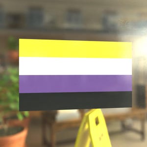May include: A rectangular sticker displaying the non-binary pride flag. The flag has horizontal stripes of yellow, white, purple, and black. The sticker is affixed to a glass surface, with a blurred background.