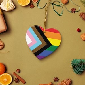May include: Heart-shaped ornament featuring the Progress Pride flag design. The ornament has a gold string for hanging. The design includes the traditional rainbow colors, along with the colors of the transgender flag, and black and brown stripes.