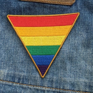 May include: A rainbow-colored triangle patch sewn onto a blue denim fabric. The patch is made of embroidered fabric and has a brown outline.