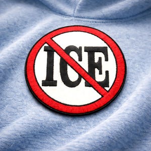 Może przedstawiać: Okrągła naszywka z czarnym napisem "ICE", przekreślonym czerwoną przekątną. Tekst znajduje się na białym tle, otoczonym czerwonym okręgiem. Naszywka jest na jasnoniebieskiej tkaninie.