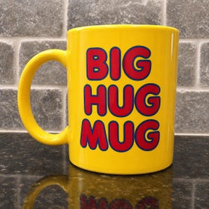 Peut inclure: Une tasse jaune vif avec une grande anse. La tasse affiche les mots "BIG HUG MUG" en lettres rouges avec des contours bleus. La tasse est posée sur une surface noire avec un fond en briques.
