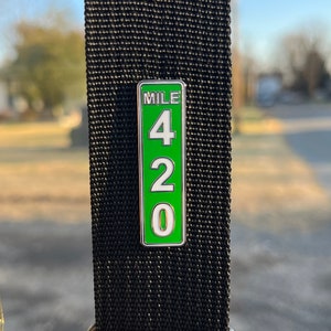 May include: A green and white enamel pin with the text "MILE 420" on a black strap. The pin is rectangular with rounded corners and a white border. The numbers are white on a green background.