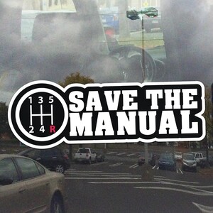 Może przedstawiać: Biała naklejka z napisem "SAVE THE MANUAL" pogrubioną czcionką i wielkimi literami. Naklejka zawiera również czarno-białą grafikę schematu zmiany biegów samochodu. Tło przedstawia parking i wnętrze samochodu.