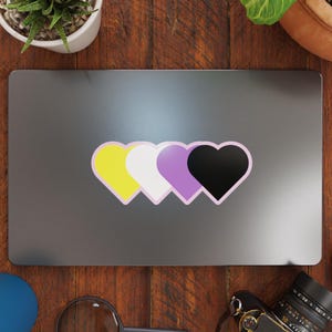 May include: A laptop computer with a sticker of three hearts in the colors of the non-binary pride flag: yellow, white, purple, and black.