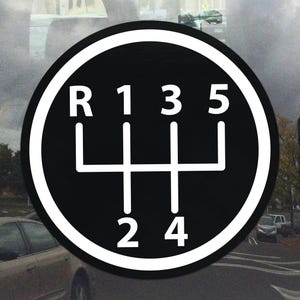 Può includere: Un adesivo in bianco e nero con lo schema del cambio di un'auto con cambio manuale. Il design circolare include la lettera "R" per la retromarcia e i numeri da 1 a 5, che indicano le posizioni delle marce. Sfondo sfocato.