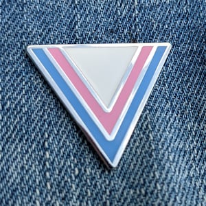 May include: A silver triangle pin with blue, pink and white enamel. The pin is in the shape of a triangle with a white centre and blue and pink stripes on the edges.