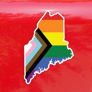 May include: A sticker of the state of Maine in the shape of the state with the colors of the Progress Pride flag. The flag has the colors red, orange, yellow, green, blue, pink, light blue, brown, and black.