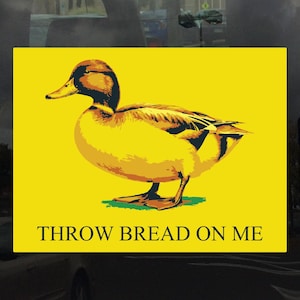 Puede incluir: Una pegatina amarilla con una imagen de dibujos animados de un pato y el texto "THROW BREAD ON ME".
