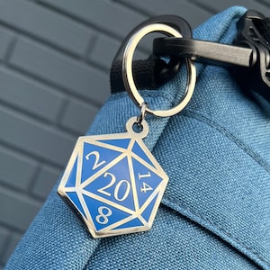 Può includere: Un portachiavi color argento con un ciondolo a dado a venti facce blu e argento. Il dado presenta i numeri 2, 8, 14 e 20. Il portachiavi è attaccato a una borsa di stoffa blu.