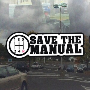 Może przedstawiać: Biało-czarna naklejka z napisem "SAVE THE MANUAL" i schematem zmiany biegów. Schemat zmiany biegów pokazuje cyfry 1, 2, 3, 4, 5 i R (wsteczny). Naklejka znajduje się na szybie samochodu.