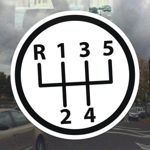 Può includere: Un adesivo bianco con bordo nero raffigurante lo schema del cambio di una trasmissione manuale. Il motivo include le lettere "R" e i numeri "1, 2, 3, 4, 5" disposti in un cerchio. Lo sfondo mostra una scena stradale.