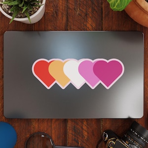 May include: A laptop sticker with four hearts in a row. The hearts are red, orange, white, and pink. The sticker is on a silver laptop.