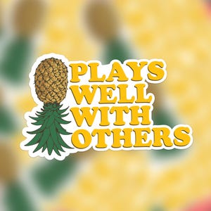 Puede incluir: Una pegatina amarilla con el texto "Plays Well With Others" en una fuente retro. Una ilustración de piña está a la izquierda del texto.