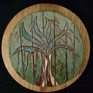 May include: A round wooden plaque with a ceramic tile depicting a stylized tree with blue-green leaves and a brown trunk. The tile is set against a green background.