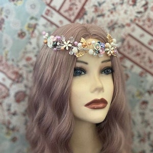 May include: A floral crown with gold leaves, white pearls, and colorful flowers. The crown is set on a mannequin head with wavy, mauve-colored hair. The mannequin is wearing a black top. The background is a floral patterned fabric.