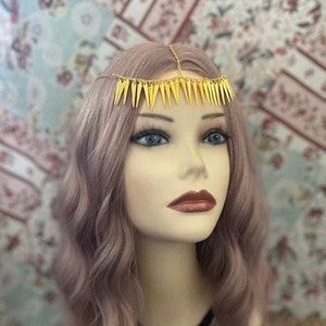 May include: A gold-colored headpiece with a chain and triangular spikes. The headpiece is worn by a person with long, wavy, light purple hair.