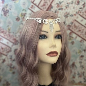 May include: A silver rhinestone headband with a dangling chain of rhinestones. The headband is worn by a person with long, wavy, light purple hair.