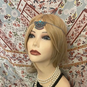 May include: A silver head chain with turquoise and red beads, worn by a mannequin with blonde hair and a pearl necklace. The mannequin is wearing a black top and is posed against a floral patterned background.
