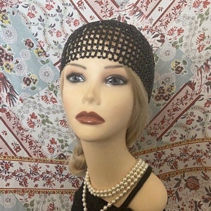 May include: A black, open-weave head covering, possibly a cap or snood, is displayed on a mannequin head. The head also features a layered pearl necklace. The background is a floral patterned fabric in shades of blue, red, and white.