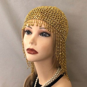 May include: A gold beaded headpiece with a netted design and beaded fringe. The headpiece is displayed on a mannequin with blonde hair, red lipstick, and a pearl necklace. The headpiece is a vintage or costume accessory.