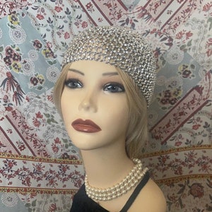 May include: A silver rhinestone headpiece. The headpiece is displayed on a mannequin with blonde hair, a pearl necklace, and a black top. The background features a floral patterned fabric.