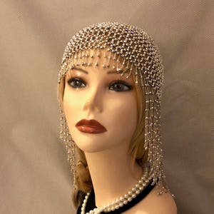 May include: A silver beaded headpiece with a net design and dangling bead strands. The headpiece covers the top of the head. The mannequin also wears a pearl necklace.