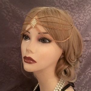 May include: Gold chain headpiece with a decorative, geometric design in the center. The headpiece is made of multiple chains that are connected together.
