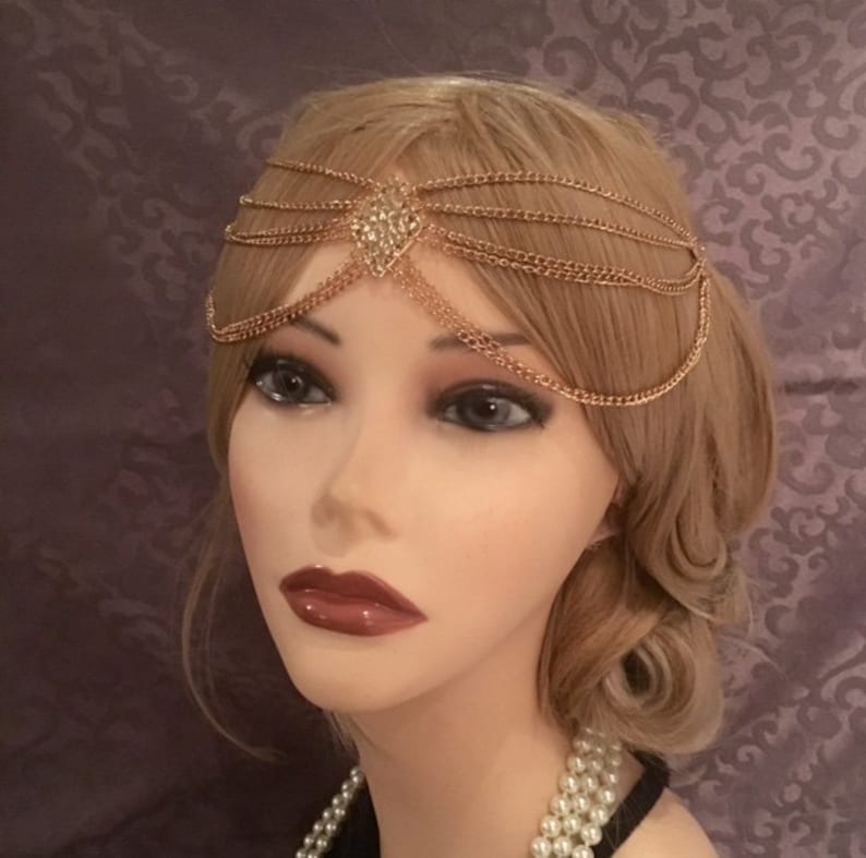 May include: Gold chain headpiece with a crystal heart design. The headpiece is made of multiple chains that are connected to form a delicate and intricate design.