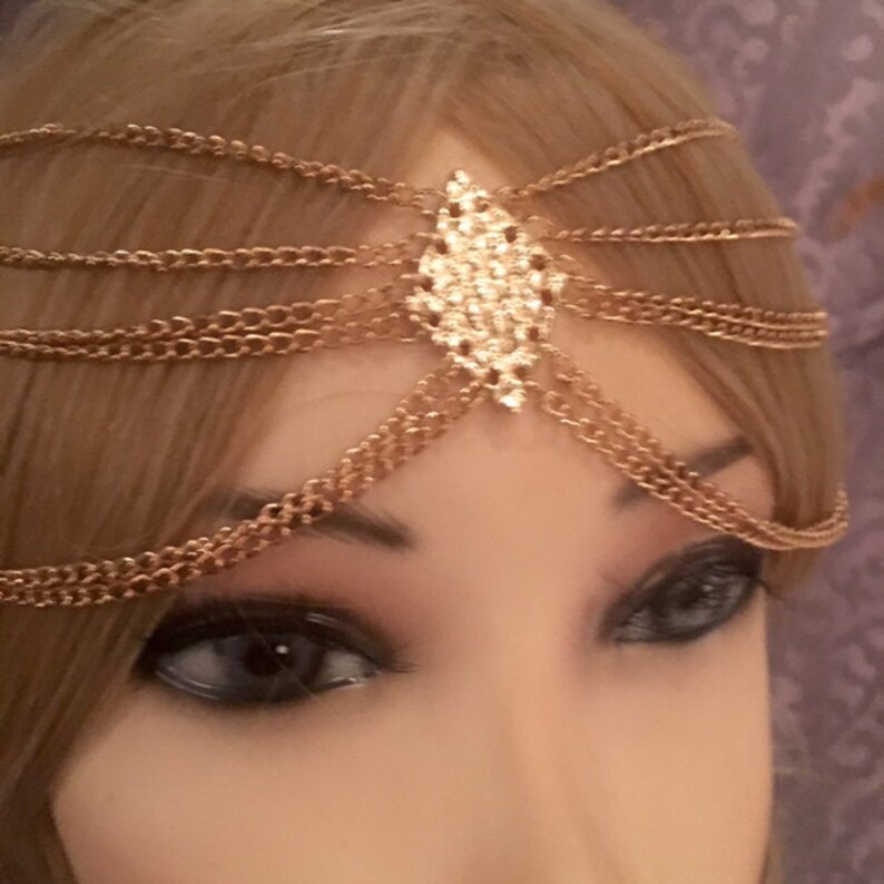 May include: Gold chain headpiece with a large, clear rhinestone centerpiece. The headpiece is made of multiple chains that are connected to form a delicate and intricate design.