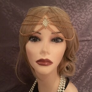 May include: Gold chain headpiece with a diamond-shaped design in the center. The headpiece is made of multiple chains that are layered on top of each other.
