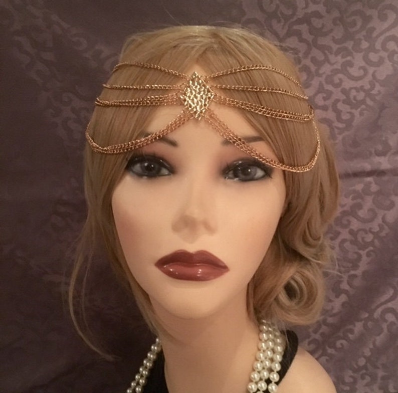 May include: Gold chain headpiece with a diamond-shaped design in the center. The headpiece is made of multiple chains that are layered on top of each other.