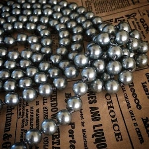 May include: A strand of gray pearl beads. The beads are round and have a shiny, metallic finish. The necklace is draped across a vintage newspaper background, adding a touch of classic elegance. The pearls are uniform in size and closely strung.